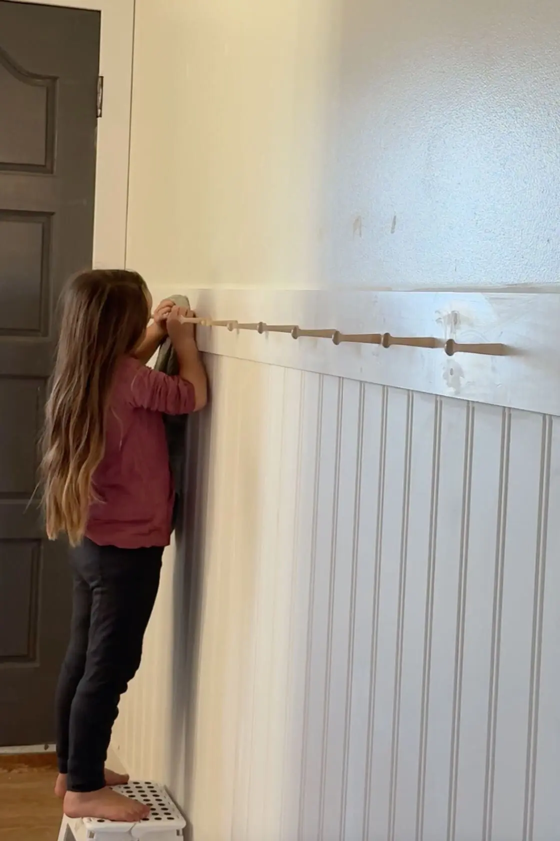 A young girl standing on a step stool, wiping down a freshly installed wooden peg rail above beadboard paneling. Her long hair and careful attention to detail emphasize the collaborative and family-oriented aspect of this DIY project.